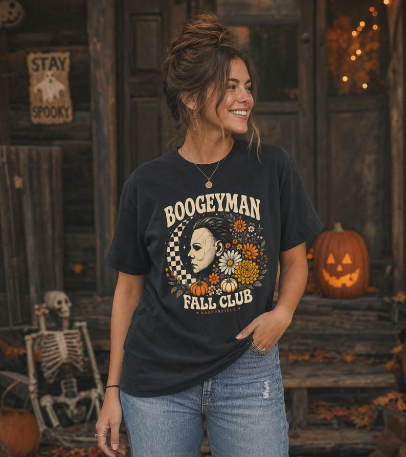 May include: A charcoal grey short-sleeve t-shirt featuring a graphic print with the text "BOOGEYMAN FALL CLUB" and "HADDONFIELD". The design includes a profile illustration of a masked figure, a checkered crescent moon, and orange flowers.