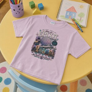 May include: A light purple short-sleeve t-shirt featuring a graphic print of the Epcot theme park. The design includes the Spaceship Earth geodesic sphere, a monorail, and various international pavilion buildings with the text This is EPCOT.