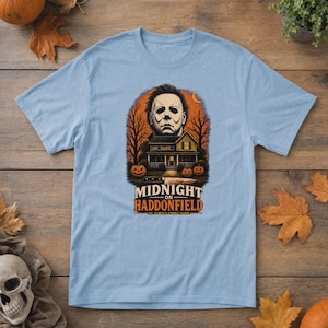 May include: A light blue t-shirt featuring a graphic print of a masked figure above a house, trees, and pumpkins. A knife is positioned below the house. The text reads MIDNIGHT ON HADDONFIELD HE ALWAYS COMES HOME in black and orange lettering.