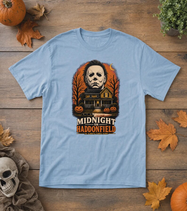 May include: A light blue t-shirt featuring a graphic print of a masked figure above a house, trees, and pumpkins. A knife is positioned below the house. The text reads MIDNIGHT ON HADDONFIELD HE ALWAYS COMES HOME in black and orange lettering.