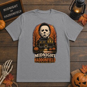 May include: A heather grey t-shirt featuring a graphic print of a masked figure above a house, pumpkins, and a knife. The text reads "MIDNIGHT ON HADDONFIELD" and "HE ALWAYS COMES HOME" in orange and white lettering against an orange background.