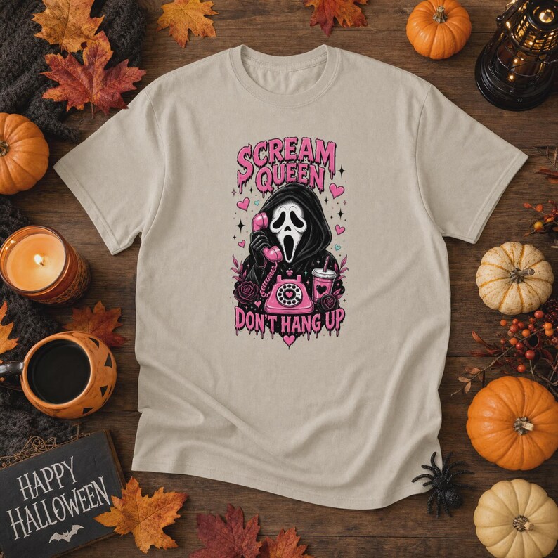 May include: A beige short-sleeve t-shirt featuring a graphic of a hooded figure in a mask holding a pink rotary telephone. The text reads "SCREAM QUEEN" and "DON'T HANG UP" in pink lettering, surrounded by pink hearts, roses, and a drink cup.