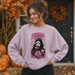 May include: A light purple crewneck sweatshirt featuring a graphic print of a hooded figure in a mask holding a pink rotary telephone. The text reads SCREAM QUEEN and DON'T HANG UP in pink lettering, surrounded by small hearts and star accents.