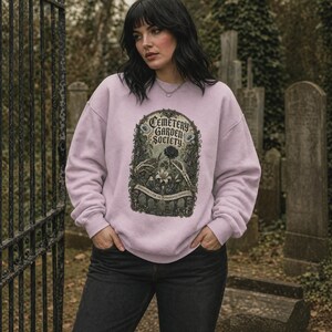 May include: A light purple crewneck sweatshirt featuring a graphic print with the text Cemetery Garden Society, Perennials for the Permanently Departed, and Open Dusk to Dawn, surrounded by skeletal hands, flowers, and gravestones.