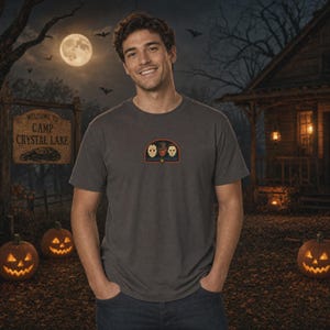 Puede incluir: Una camiseta de manga corta de color gris carb&oacute;n con un peque&ntilde;o gr&aacute;fico centrado en el pecho. El gr&aacute;fico es un emblema arqueado que contiene tres m&aacute;scaras de personajes de pel&iacute;culas de terror dispuestas horizontalmente, enmarcadas por finas l&iacute;neas naranjas y azules.