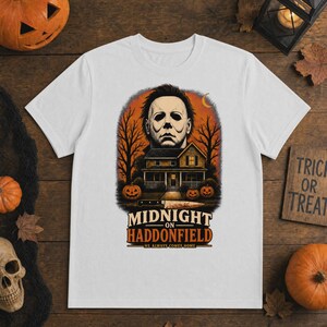 May include: A white short-sleeve t-shirt featuring a graphic of a masked figure above a house, trees, and pumpkins. The text reads MIDNIGHT ON HADDONFIELD HE ALWAYS COMES HOME. A knife is positioned horizontally below the house illustration.