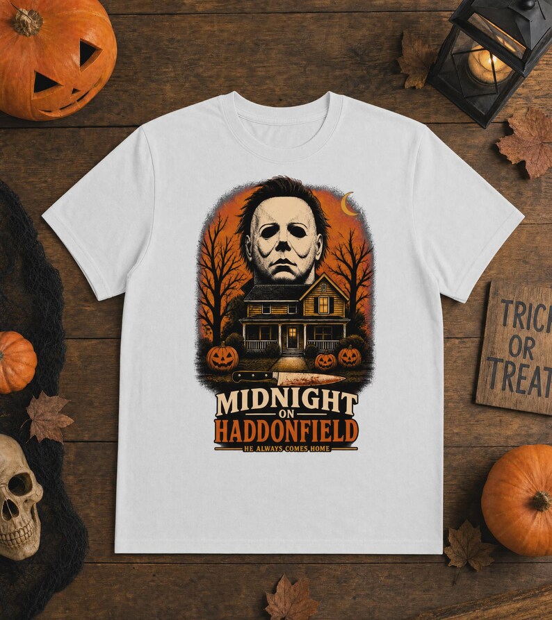 May include: A white short-sleeve t-shirt featuring a graphic of a masked figure above a house, trees, and pumpkins. The text reads MIDNIGHT ON HADDONFIELD HE ALWAYS COMES HOME. A knife is positioned horizontally below the house illustration.