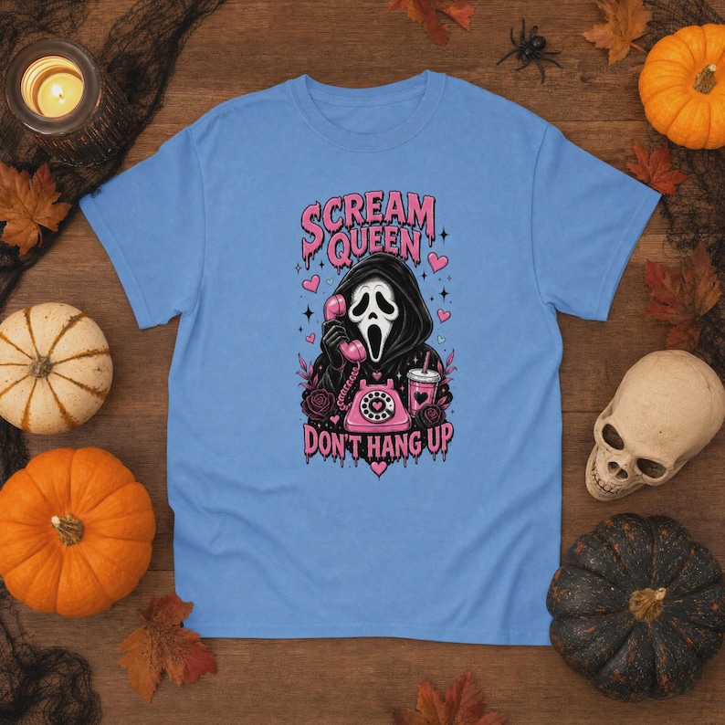 May include: A light blue t-shirt featuring a graphic of a hooded figure in a mask holding a pink rotary telephone. The text reads SCREAM QUEEN DON'T HANG UP in pink letters. The design includes pink hearts, a pink drink cup, and dark roses.