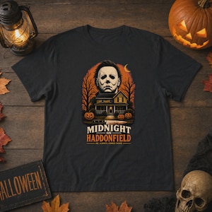 May include: A black t-shirt featuring a graphic of a masked figure above a house, trees, and pumpkins. A knife is positioned below the house. The text reads MIDNIGHT ON HADDONFIELD HE ALWAYS COMES HOME in white and orange lettering on the front.