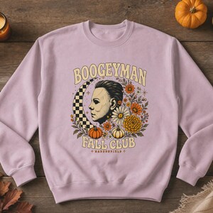 May include: A light purple crewneck sweatshirt featuring a graphic print of a masked face, a checkered crescent moon, orange and white pumpkins, and various flowers. The text reads BOOGEYMAN FALL CLUB and HADDONFIELD in a curved font style.