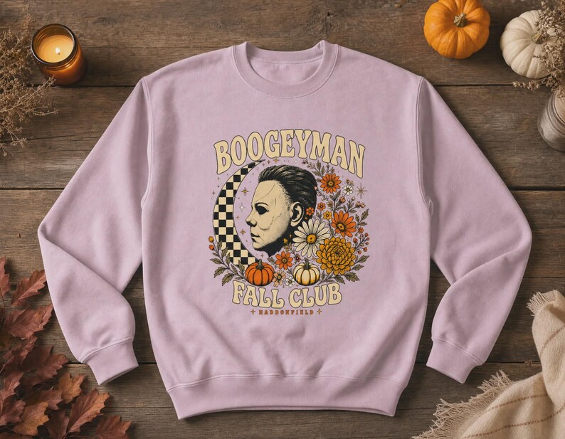 May include: A light purple crewneck sweatshirt featuring a graphic print of a masked face, a checkered crescent moon, orange and white pumpkins, and various flowers. The text reads BOOGEYMAN FALL CLUB and HADDONFIELD in a curved font style.