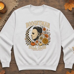 May include: A white crewneck sweatshirt featuring a graphic print of a masked figure in profile, a checkered crescent moon, autumn flowers, and pumpkins. The text reads BOOGEYMAN FALL CLUB and HADDONFIELD in a tan, arched, retro-style font.