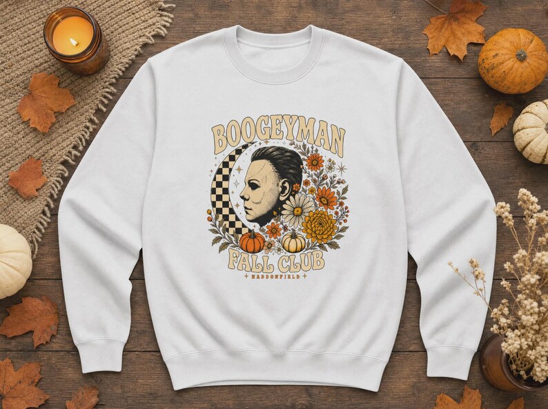May include: A white crewneck sweatshirt featuring a graphic print of a masked figure in profile, a checkered crescent moon, autumn flowers, and pumpkins. The text reads BOOGEYMAN FALL CLUB and HADDONFIELD in a tan, arched, retro-style font.