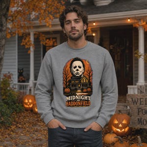 May include: A heather grey crewneck sweatshirt featuring a graphic print of a masked figure, a house, and pumpkins. The text reads MIDNIGHT ON HADDONFIELD and HE ALWAYS COMES HOME. The design includes orange and black tones with bare trees.