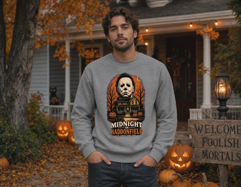 May include: A heather grey crewneck sweatshirt featuring a graphic print of a masked figure, a house, and pumpkins. The text reads MIDNIGHT ON HADDONFIELD and HE ALWAYS COMES HOME. The design includes orange and black tones with bare trees.