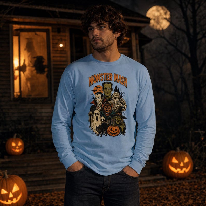 May include: A light blue long-sleeve shirt featuring a graphic print with the text MONSTER MASH in orange letters. The illustration depicts various classic horror characters including a vampire, Frankenstein's monster, a mummy, a werewolf, a ghost, a witch, a black cat, and a jack-o'-lantern.