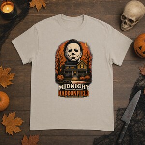 May include: A beige short-sleeve t-shirt featuring a graphic print of a masked figure above a house, trees, and pumpkins. The text reads "MIDNIGHT ON HADDONFIELD" and "HE ALWAYS COMES HOME" with a knife illustration below the house.