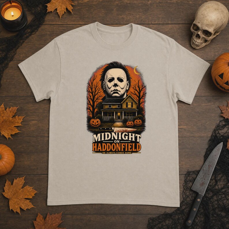 May include: A beige short-sleeve t-shirt featuring a graphic print of a masked figure above a house, trees, and pumpkins. The text reads "MIDNIGHT ON HADDONFIELD" and "HE ALWAYS COMES HOME" with a knife illustration below the house.