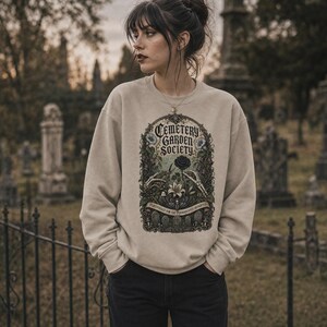 May include: A beige crewneck sweatshirt featuring a detailed graphic print. The design displays the text Cemetery Garden Society, skeletal hands, a black rose, a white lily, and gravestones within an ornate, dark, botanical frame.