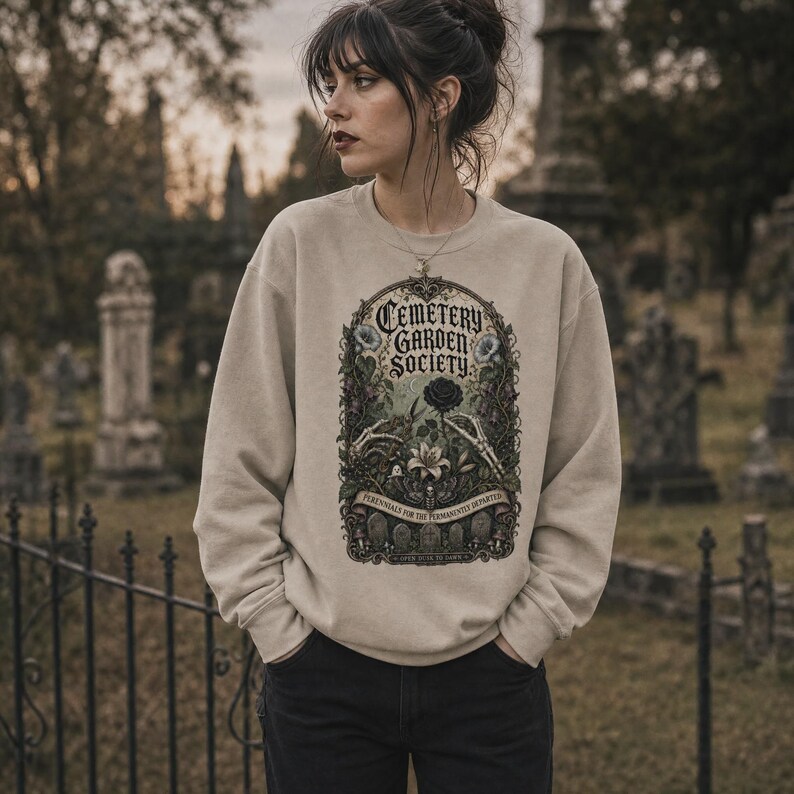 May include: A beige crewneck sweatshirt featuring a detailed graphic print. The design displays the text Cemetery Garden Society, skeletal hands, a black rose, a white lily, and gravestones within an ornate, dark, botanical frame.