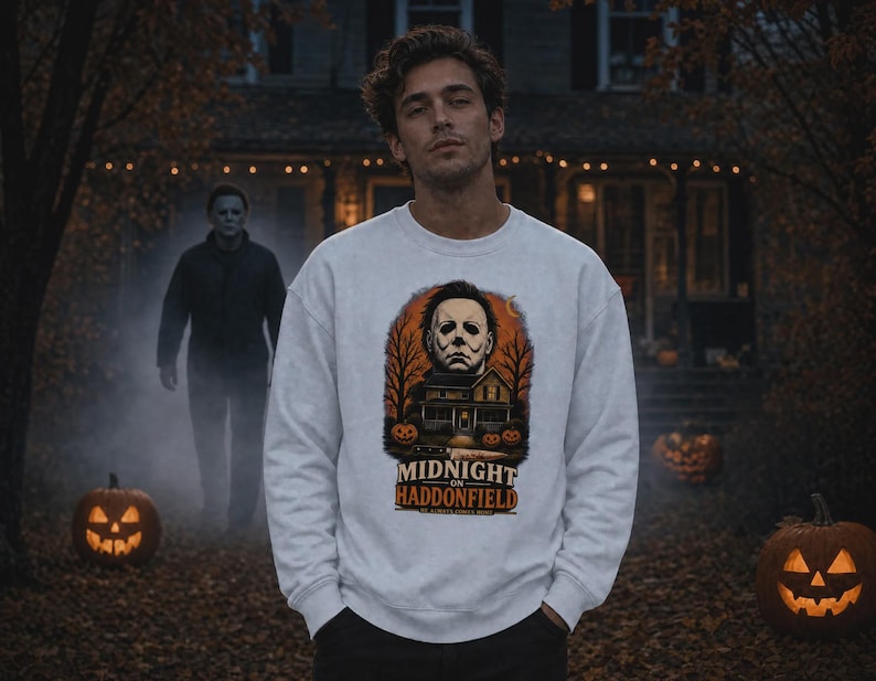 May include: A light grey crewneck sweatshirt featuring a graphic print of a masked figure above a house, two jack-o'-lanterns, and a knife. The text reads MIDNIGHT ON HADDONFIELD and HE ALWAYS COMES HOME in orange and white lettering.