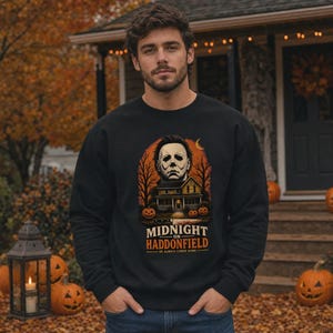 May include: A black long-sleeve sweatshirt featuring a graphic print of a masked figure above a house, surrounded by trees and pumpkins. The text reads MIDNIGHT ON HADDONFIELD HE ALWAYS COMES HOME. The design includes an orange crescent moon.