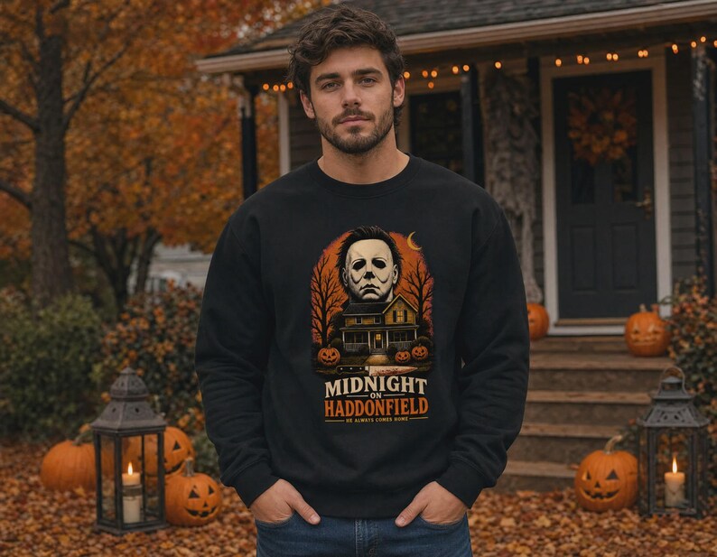 May include: A black long-sleeve sweatshirt featuring a graphic print of a masked figure above a house, surrounded by trees and pumpkins. The text reads MIDNIGHT ON HADDONFIELD HE ALWAYS COMES HOME. The design includes an orange crescent moon.