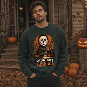 May include: A dark green long-sleeve sweatshirt featuring a graphic print of a masked character, a house, and pumpkins. The text reads MIDNIGHT ON HADDONFIELD HE ALWAYS COMES HOME. The design includes bare trees and an orange moon background.