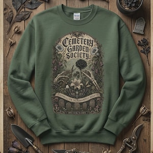 May include: An olive green crewneck sweatshirt featuring a graphic print with the text Cemetery Garden Society, Perennials for the Permanently Departed, and Open Dusk to Dawn, depicting skeleton hands, a black rose, a moth, and tombstones.