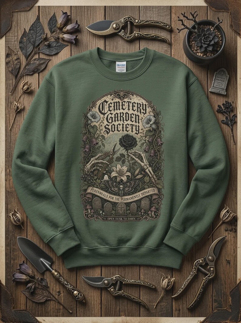 May include: An olive green crewneck sweatshirt featuring a graphic print with the text Cemetery Garden Society, Perennials for the Permanently Departed, and Open Dusk to Dawn, depicting skeleton hands, a black rose, a moth, and tombstones.