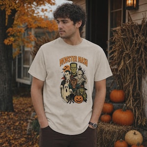 May include: A beige short-sleeve t-shirt featuring a graphic print with the text MONSTER MASH in orange letters. The illustration depicts various Halloween characters including a vampire, Frankenstein's monster, a mummy, a werewolf, a ghost, a witch, a black cat, and a jack-o'-lantern.
