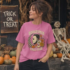 May include: A pink short-sleeve t-shirt featuring a graphic print of a masked figure in profile, a checkered crescent moon, pumpkins, and various flowers. The text reads BOOGEYMAN FALL CLUB in a curved font above and below the central illustration.