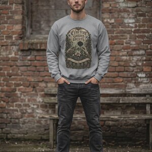 May include: A light grey crewneck sweatshirt featuring a graphic print with the text "CEMETERY GARDEN SOCIETY" in a decorative frame, surrounded by illustrations of skeletal hands, flowers, and tombstones, paired with dark grey denim jeans.