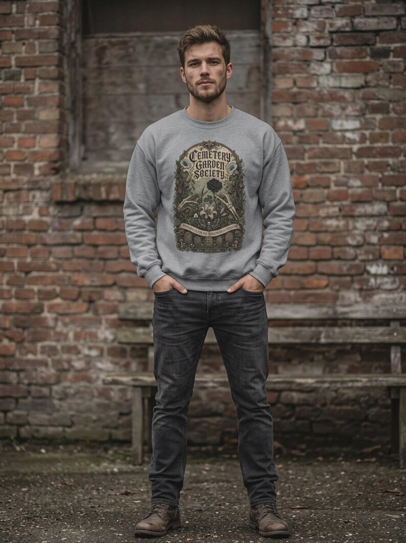 May include: A light grey crewneck sweatshirt featuring a graphic print with the text "CEMETERY GARDEN SOCIETY" in a decorative frame, surrounded by illustrations of skeletal hands, flowers, and tombstones, paired with dark grey denim jeans.