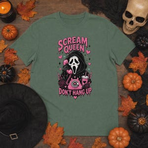 May include: A sage green t-shirt featuring a graphic of a hooded figure in a mask holding a pink rotary telephone. The text reads "SCREAM QUEEN" and "DON'T HANG UP" in pink. The design includes pink hearts, a pink drink cup, and roses.