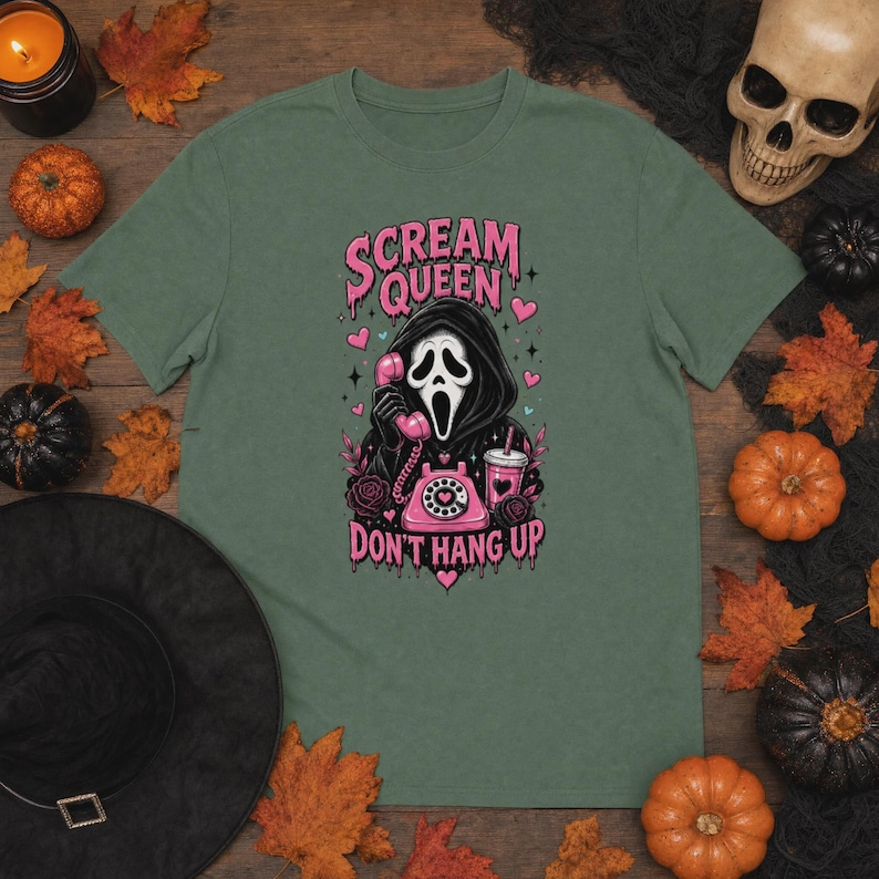 May include: A sage green t-shirt featuring a graphic of a hooded figure in a mask holding a pink rotary telephone. The text reads "SCREAM QUEEN" and "DON'T HANG UP" in pink. The design includes pink hearts, a pink drink cup, and roses.