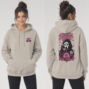 May include: A beige hooded sweatshirt featuring a small pink rotary phone graphic on the front left chest. The back displays a large graphic of a masked figure holding a pink rotary phone with the text Scream Queen Don't Hang Up in pink.