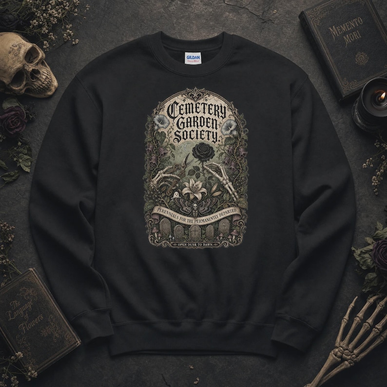 May include: A black crewneck sweatshirt featuring a graphic print with the text Cemetery Garden Society, Perennials for the Permanently Departed, and Open Dusk to Dawn. The design includes skeleton hands, a black rose, a moth, and tombstones.