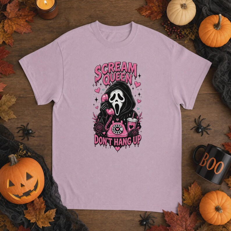 May include: A light purple t-shirt featuring a graphic of a hooded figure in a mask holding a pink rotary telephone. The text reads "SCREAM QUEEN" and "DON'T HANG UP" in a dripping pink font, surrounded by small hearts, roses, and a drink cup.