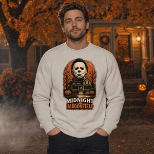 May include: A beige crewneck sweatshirt featuring a graphic print of a masked figure above a house, bare trees, and pumpkins. The text reads MIDNIGHT ON HADDONFIELD and HE ALWAYS COMES HOME. A knife is positioned horizontally below the house illustration.