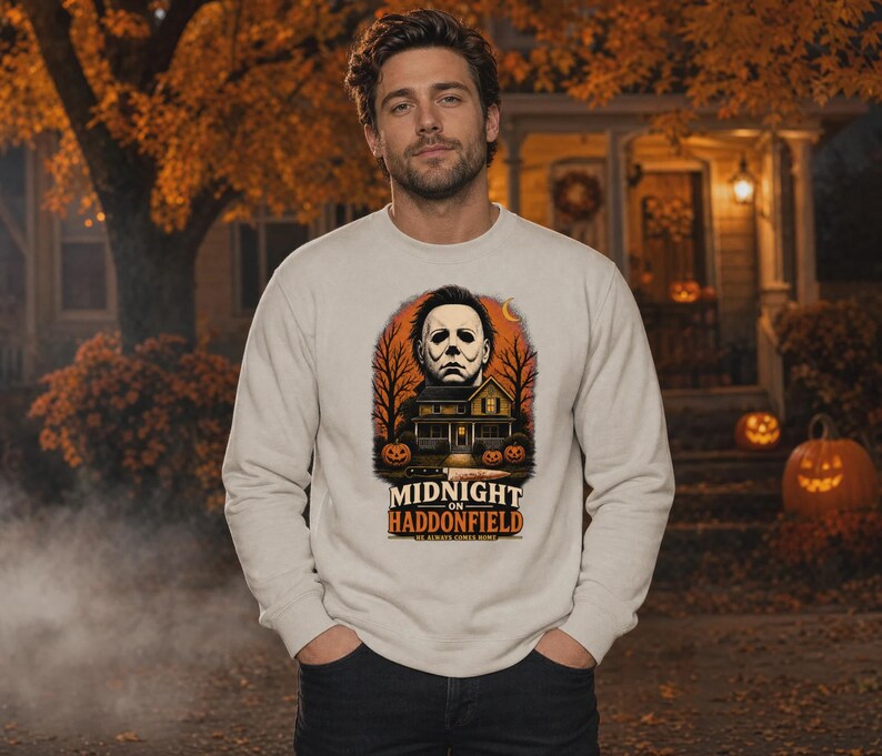 May include: A beige crewneck sweatshirt featuring a graphic print of a masked figure above a house, bare trees, and pumpkins. The text reads MIDNIGHT ON HADDONFIELD and HE ALWAYS COMES HOME. A knife is positioned horizontally below the house illustration.