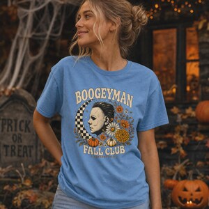 May include: A light blue short-sleeve t-shirt featuring a graphic print of a masked face in profile, a checkered crescent moon, orange and white pumpkins, and various autumn flowers, with the text BOOGEYMAN FALL CLUB printed in cream.