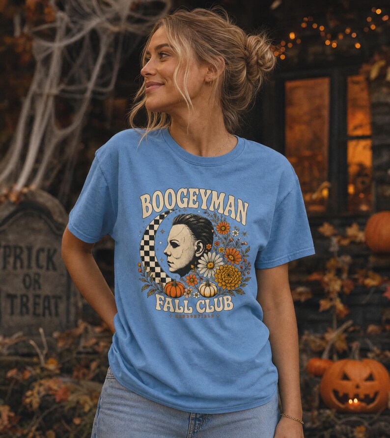 May include: A light blue short-sleeve t-shirt featuring a graphic print of a masked face in profile, a checkered crescent moon, orange and white pumpkins, and various autumn flowers, with the text BOOGEYMAN FALL CLUB printed in cream.