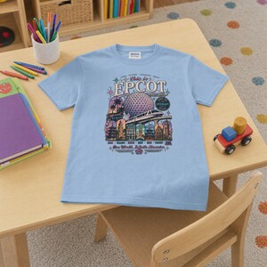 May include: A light blue short-sleeve t-shirt featuring a graphic print of the Epcot theme park. The design includes the Spaceship Earth structure, a monorail, and various international pavilion landmarks with the text This is EPCOT.