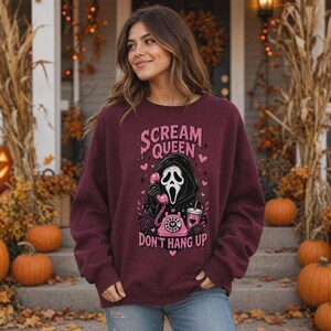 May include: A maroon crewneck sweatshirt featuring a graphic print of a masked figure holding a pink rotary telephone. The text reads "SCREAM QUEEN" and "DON'T HANG UP" in pink lettering, surrounded by small hearts, stars, and a pink drink cup.