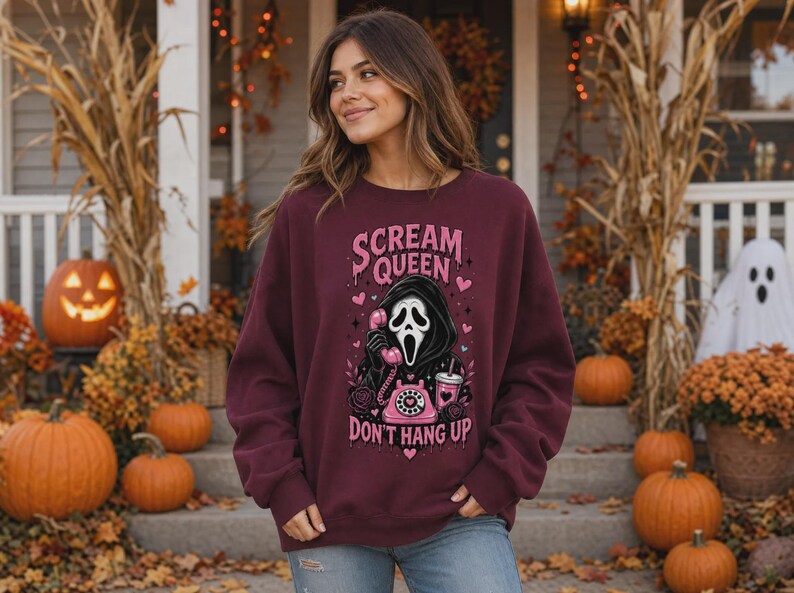 May include: A maroon crewneck sweatshirt featuring a graphic print of a masked figure holding a pink rotary telephone. The text reads "SCREAM QUEEN" and "DON'T HANG UP" in pink lettering, surrounded by small hearts, stars, and a pink drink cup.