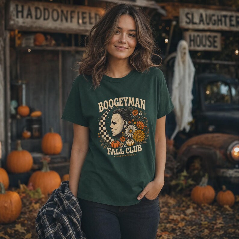 May include: A dark green short-sleeved t-shirt featuring a graphic print with the text BOOGEYMAN FALL CLUB, a profile illustration of a masked figure, a checkered crescent moon, and various orange and white flowers and pumpkins.