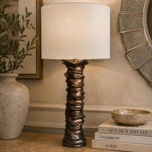 May include: A table lamp with a cylindrical white fabric shade and a tall, slender base featuring a dark bronze finish with a rippled, textured surface that mimics the appearance of draped fabric or organic, flowing vertical folds.