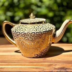 May include: A cream-colored ceramic teapot featuring a continuous, intricate gold-colored squiggly line pattern covering the body and lid. The teapot includes a gold-trimmed handle, a gold-trimmed spout, and a matching lid with a gold-colored finial.