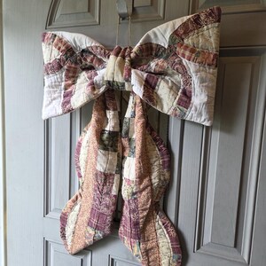 Könnte beinhalten: Eine große dekorative Schleife aus gestepptem Stoff mit einem Patchwork-Design in den Farben Burgunderrot, Creme, Beige und gedämpftem Grün. Die Schleife hat zwei breite Schlaufen und zwei lange, herabhängende Enden mit komplexen Steppmustern.